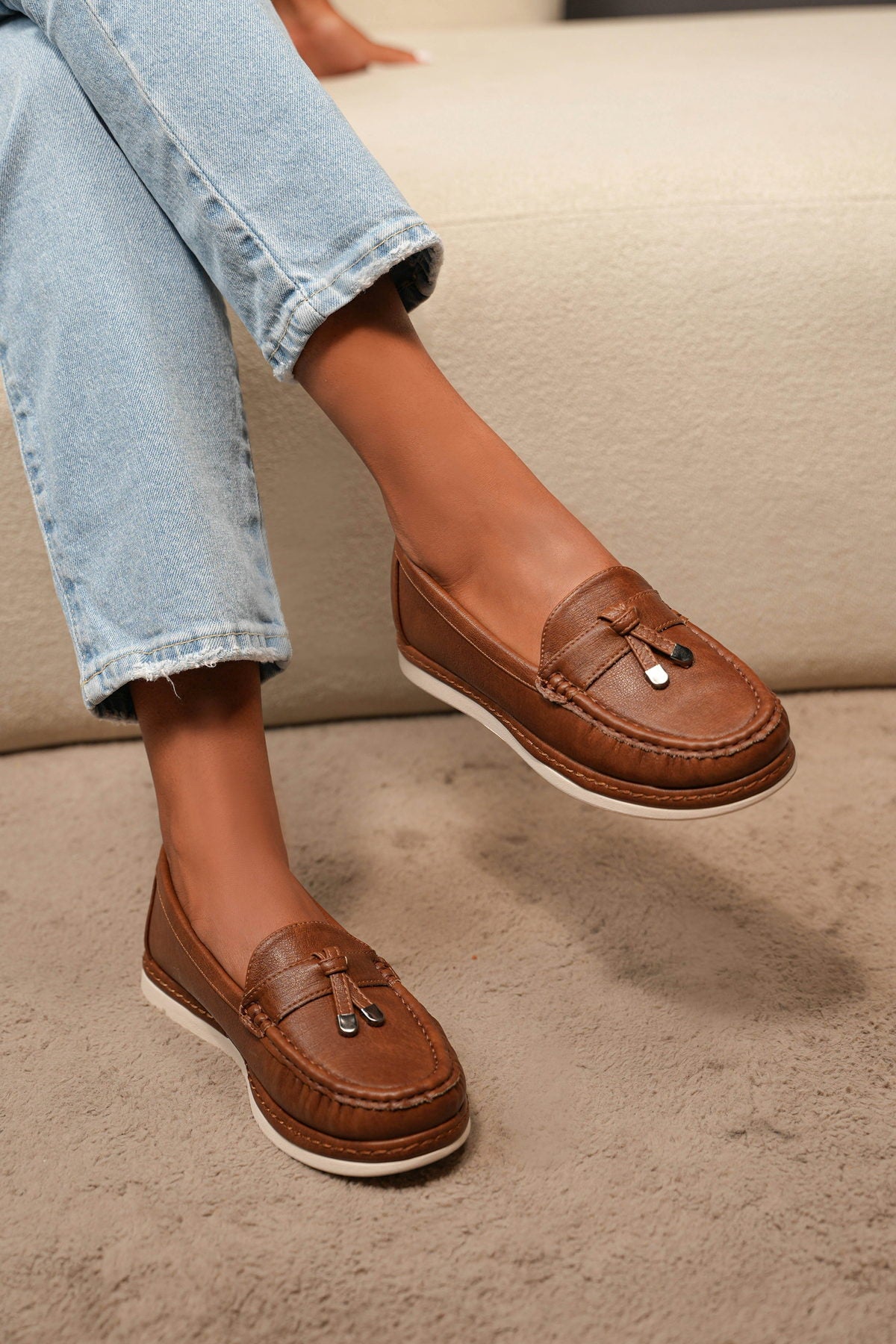 Brown leather tassel loafers worn with light blue jeans on a cream couch